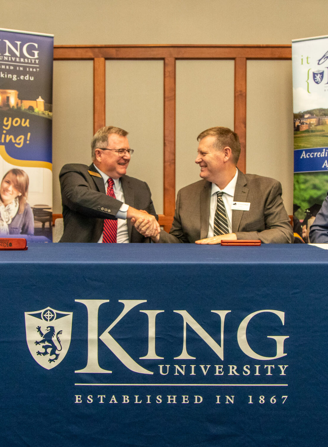Two men in suits smile and shake hands while seated at a table covered with a blue King University cloth. Banners and a sign with “King University Established in 1867” are visible in the background.