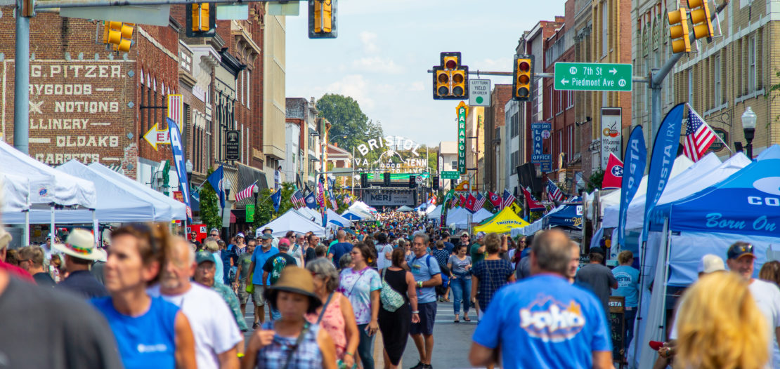 A lively street festival with crowds walking among vendor tents, under traffic lights and banners reading “Bristol,” surrounded by historic brick buildings and American flags, on a sunny day.