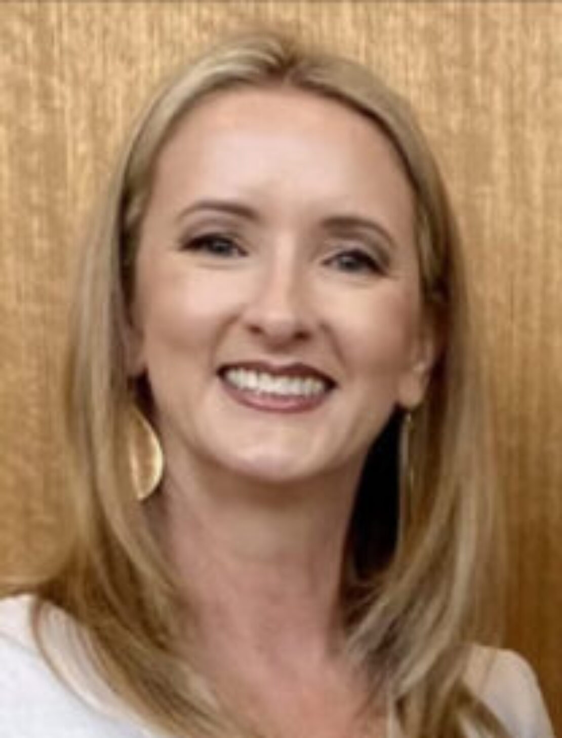 A woman with straight blonde hair, wearing a white top and large gold hoop earrings, smiles in front of a light brown wooden background.