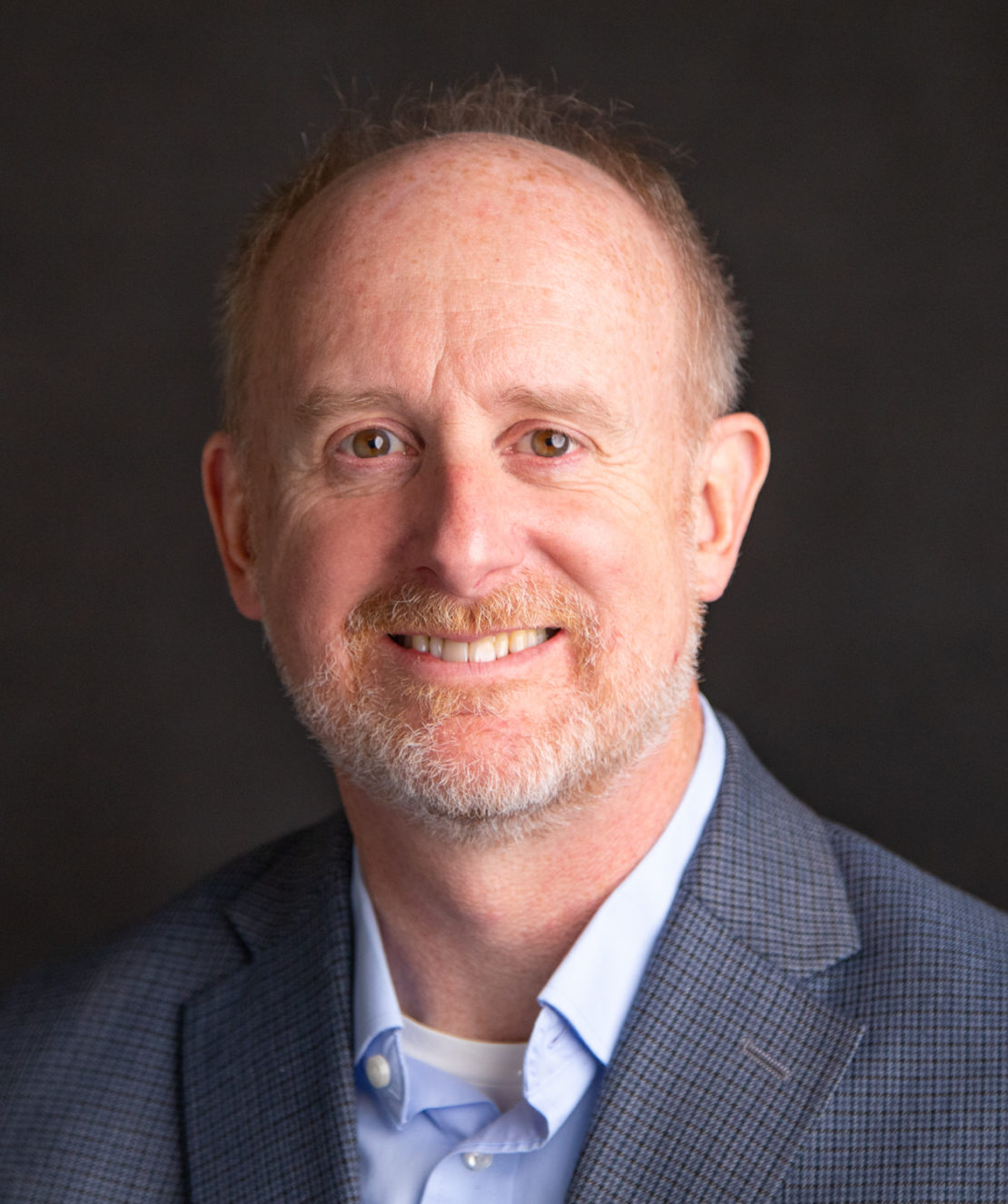 A middle-aged man with short reddish-blond hair, light beard, and wearing a gray checked blazer over a light blue shirt, smiling in front of a dark background.