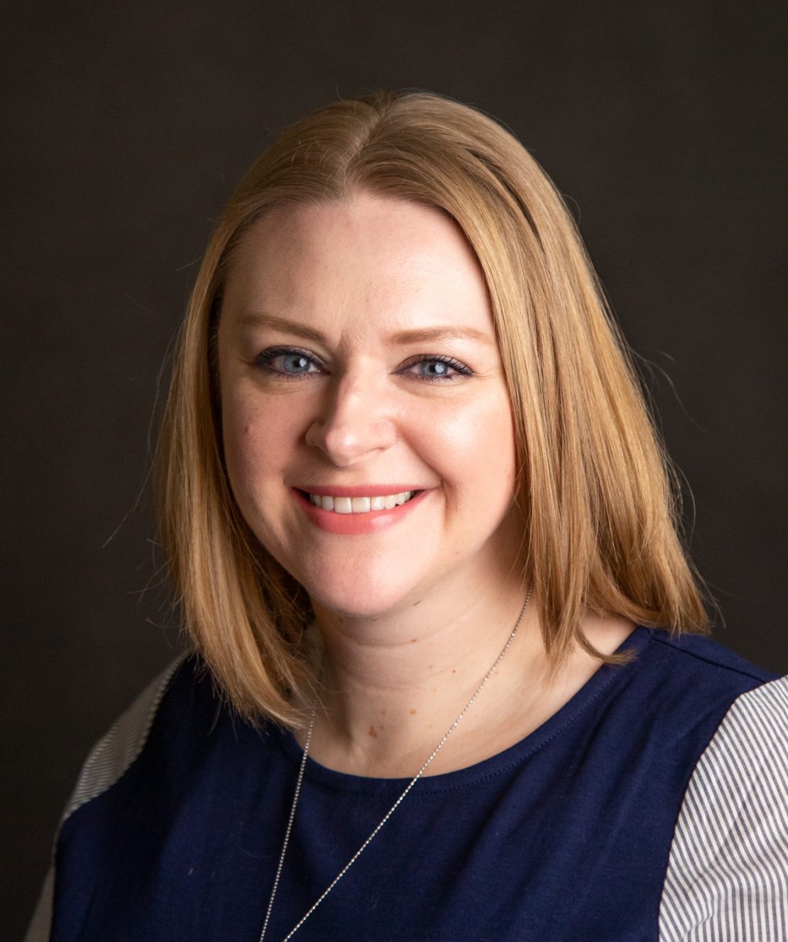A woman with straight, shoulder-length blonde hair smiles at the camera. She is wearing a navy blue top with light gray sleeves and a necklace, with a dark, plain background.