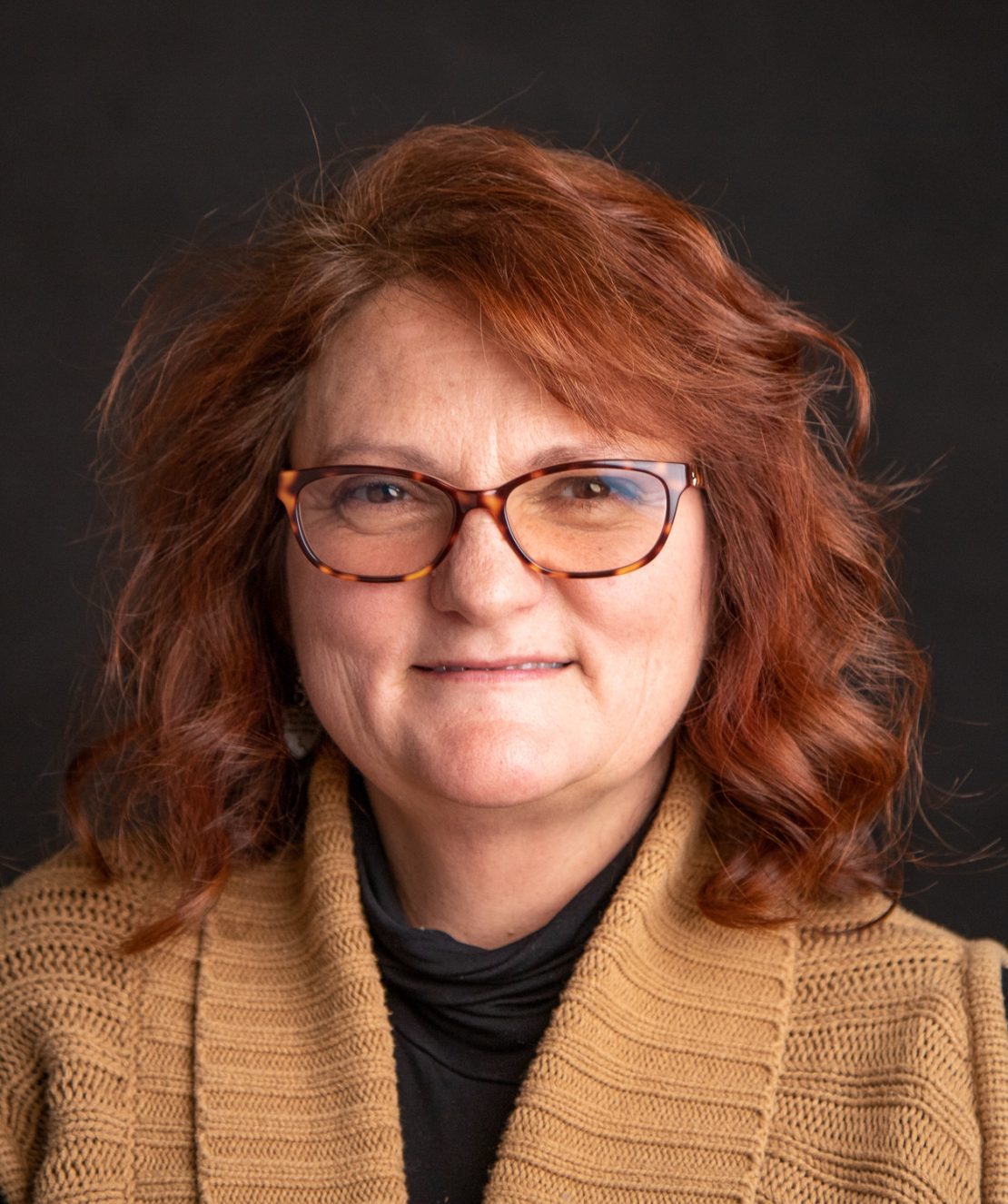 A woman with wavy red hair and glasses, wearing a brown knit cardigan over a black top, poses in front of a dark background, smiling slightly.