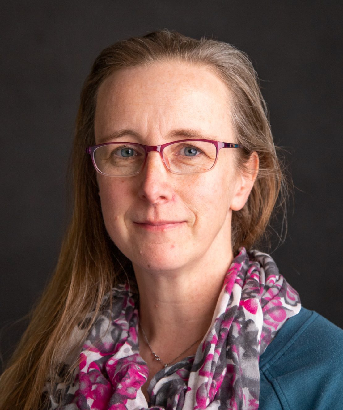 A woman with long light brown hair, wearing glasses and a colorful floral scarf, looks at the camera with a slight smile against a dark background.
