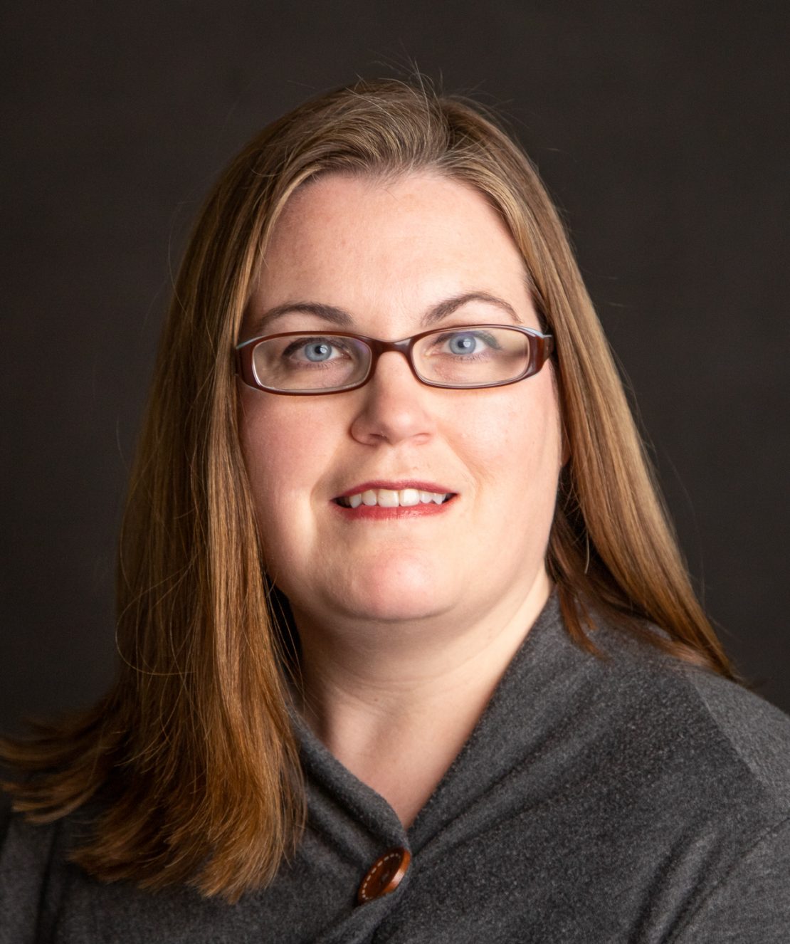 A woman with long, light brown hair and glasses is wearing a gray top and smiling slightly. She is posed in front of a dark, neutral background.