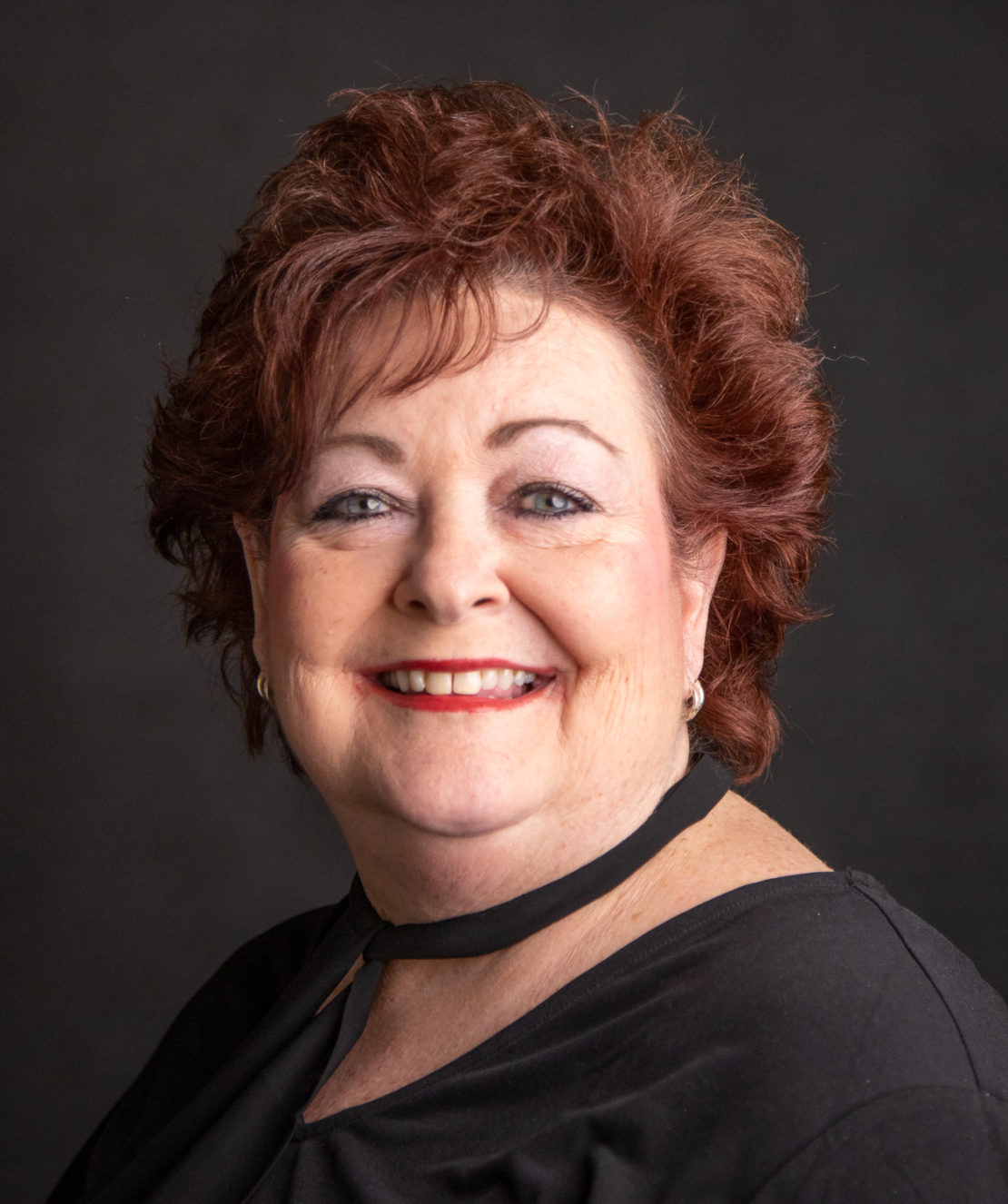 A smiling older woman with short, curly auburn hair and light makeup, wearing a black top against a dark background.