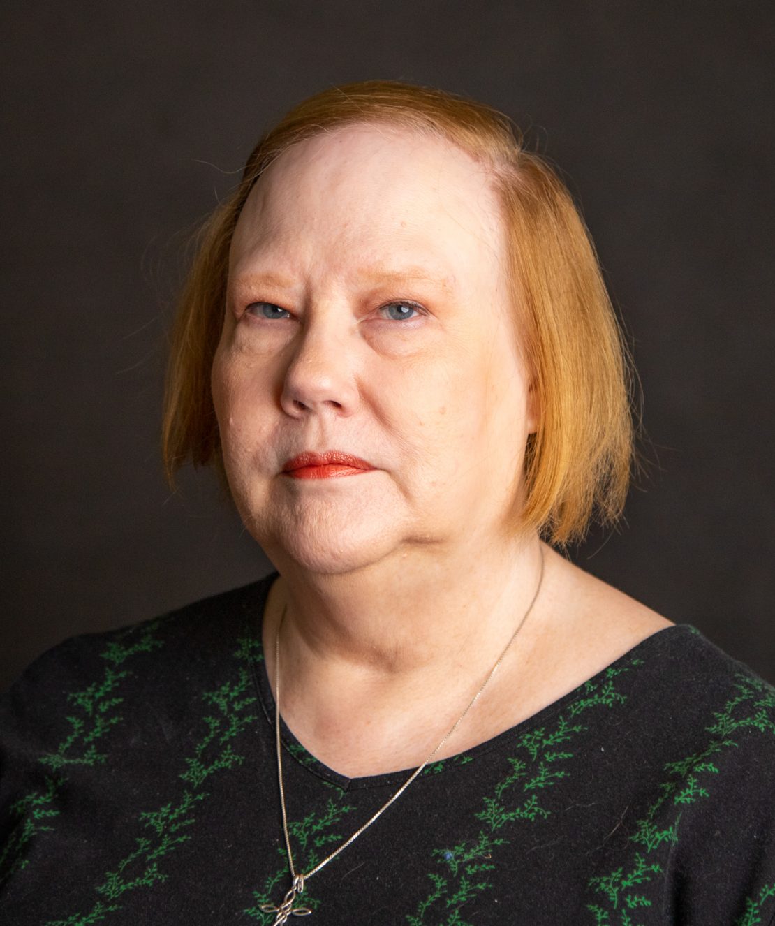 A woman with short, light brown hair and fair skin, wearing a black top with green patterns and a silver necklace, poses against a dark background with a neutral expression.