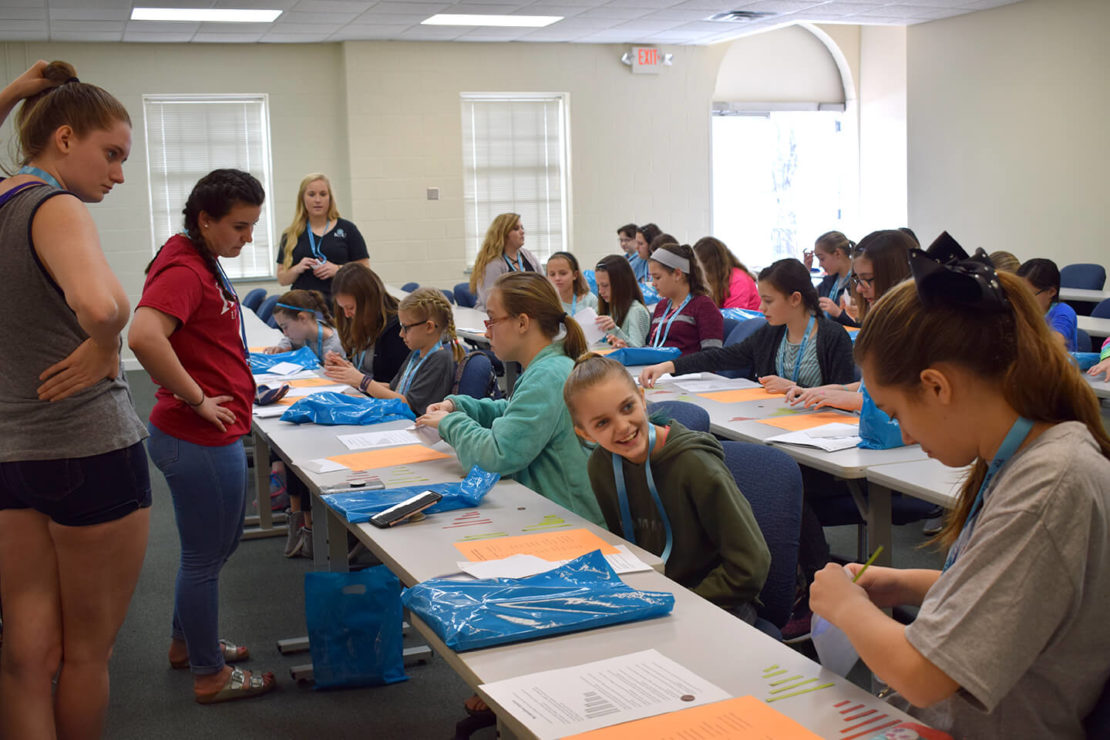 A classroom with students sitting at desks working on papers, some talking and smiling. Two women stand at the front and side of the room, overseeing the group. Bright natural light comes through large windows.