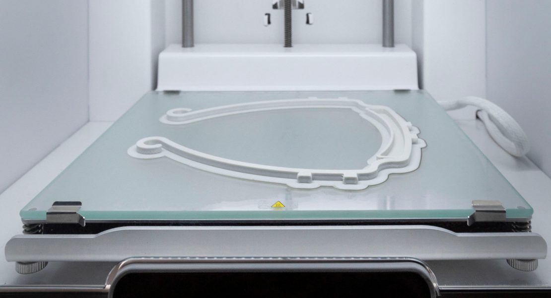 A 3D printer is printing a curved, white plastic object on a glass build plate inside the printers enclosure. The partially completed print appears to be some sort of frame or support structure.