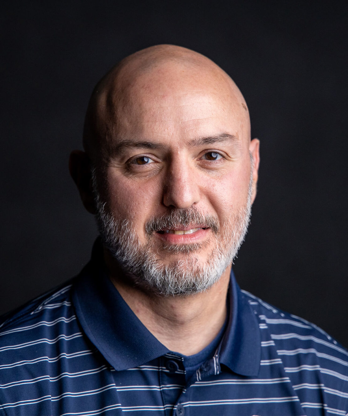 A middle-aged man with a shaved head and gray beard, wearing a blue polo shirt with white stripes, poses in front of a dark background, smiling softly.