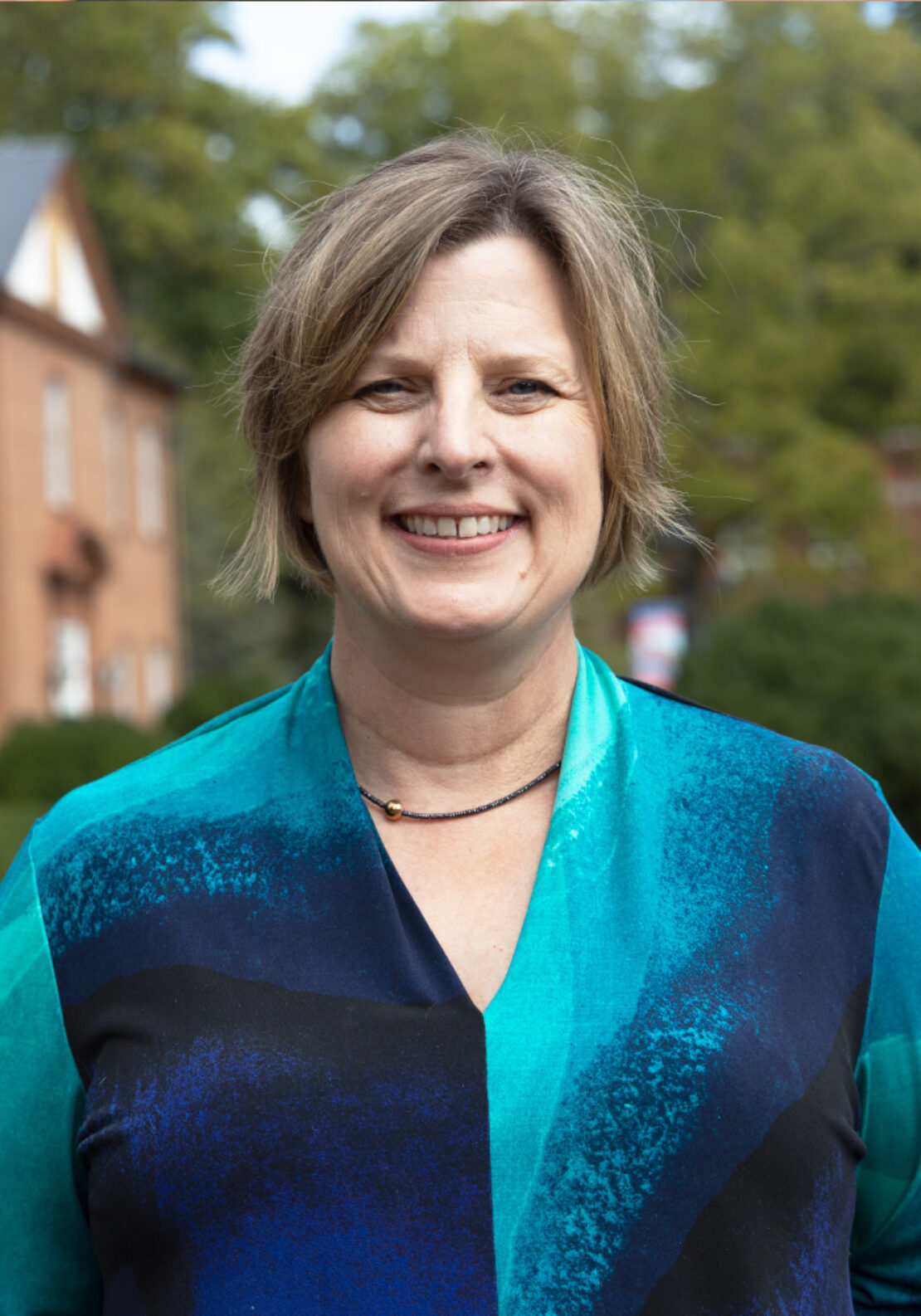 A woman with short, light brown hair smiles outdoors. She is wearing a blue and teal patterned blouse and a black necklace, with trees and a red-brick building blurred in the background.