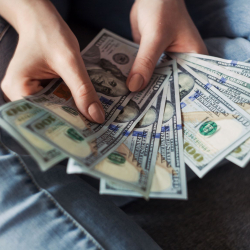 A person wearing jeans holds and fans out several $100 bills in both hands, displaying the money.
