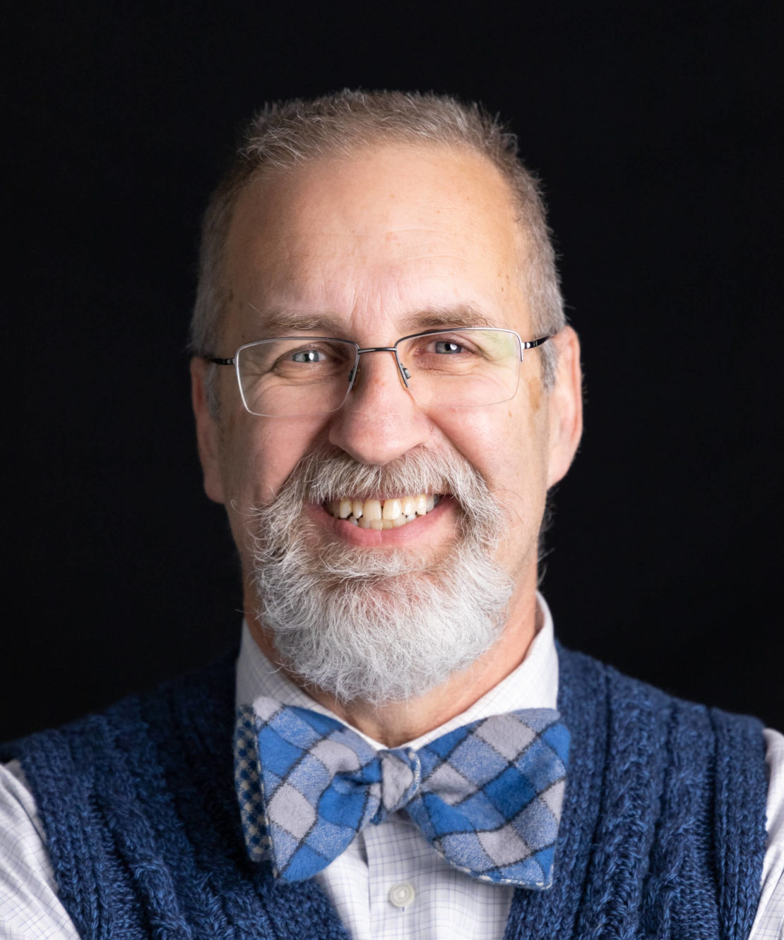 A smiling older man with gray hair, a beard, and glasses, wearing a blue patterned bow tie, a white shirt, and a navy blue sweater vest, poses against a black background.