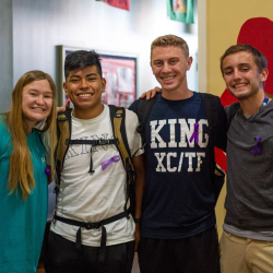 Four smiling young adults stand close together indoors, each wearing a purple awareness ribbon. Two have backpacks, one wears a KING XC/TF shirt, and they are in a brightly lit room with colorful decorations.