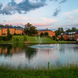 A tranquil pond with a fountain is surrounded by grassy slopes and trees, with large brick buildings in the background under a partly cloudy evening sky.
