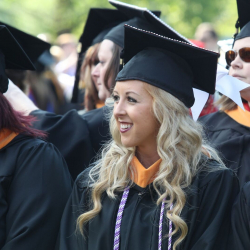 A smiling woman with long blonde hair in a black graduation gown and cap stands among other graduates at an outdoor ceremony. She wears an honor cord and looks to the side.