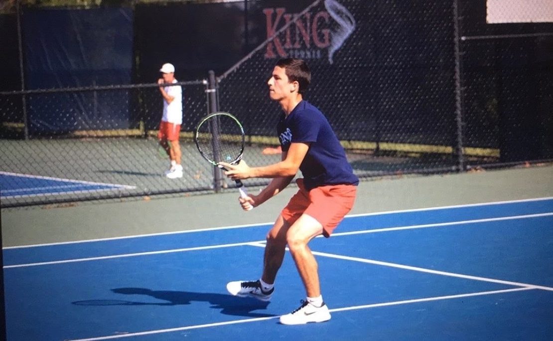 A young man in a navy shirt and red shorts prepares to hit a tennis ball on an outdoor court. Another player stands in the background. Both are holding rackets. The court is blue with a black chain-link fence behind them.