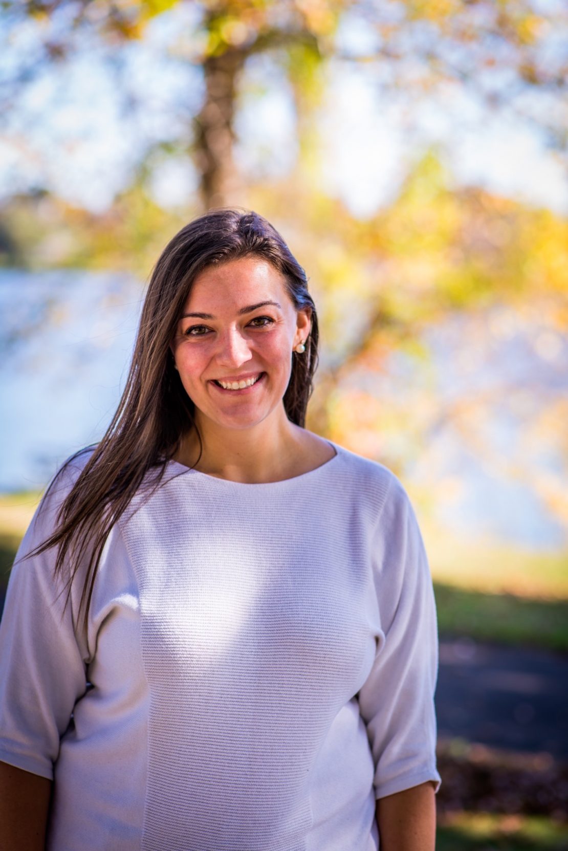 A woman with long brown hair, wearing a white top, smiles outdoors. The background shows blurred autumn trees with yellow leaves and a body of water in soft sunlight.