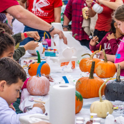 Children and adults sit around a table painting pumpkins at an outdoor event. The table is covered with art supplies, paint, and paper towels, and the participants are focused on decorating their pumpkins.