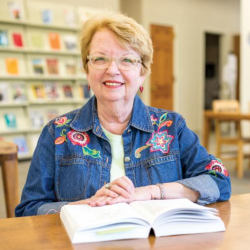 Smiling older woman with glasses and short blond hair sits at a table with an open book. She wears a denim jacket with colorful floral embroidery. Bookshelves are visible in the background.