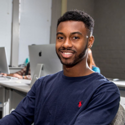A young man with short hair and a beard, wearing a navy blue long-sleeve shirt, sits smiling in a computer lab with Apple desktops in the background.