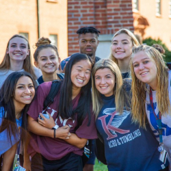 A group of eight smiling teenagers poses together outdoors in front of a brick building on a sunny day. Most wear casual clothing and have lanyards or backpacks.