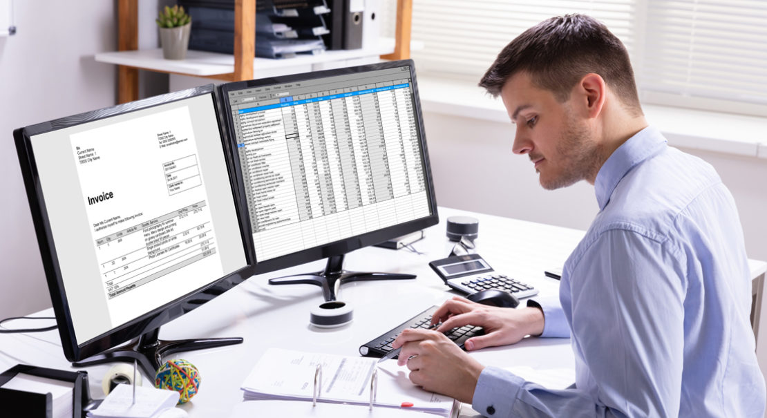 A man works at a desk with two monitors; one displays an invoice, and the other shows a spreadsheet. He uses a calculator, surrounded by papers, a keyboard, and office supplies in a well-lit office.