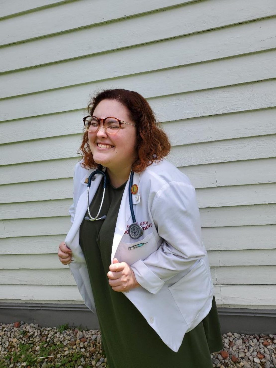 A person wearing a white medical coat and stethoscope smiles broadly while standing outside in front of a white wooden wall. They have glasses, curly hair, and are holding the sides of their coat.