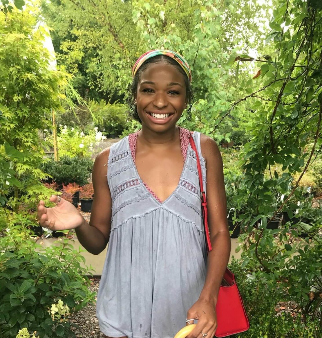 A smiling woman stands outdoors surrounded by lush green plants. She is wearing a light gray sleeveless top, a colorful headband, and carries a bright red bag. Sunlight filters through the trees behind her.