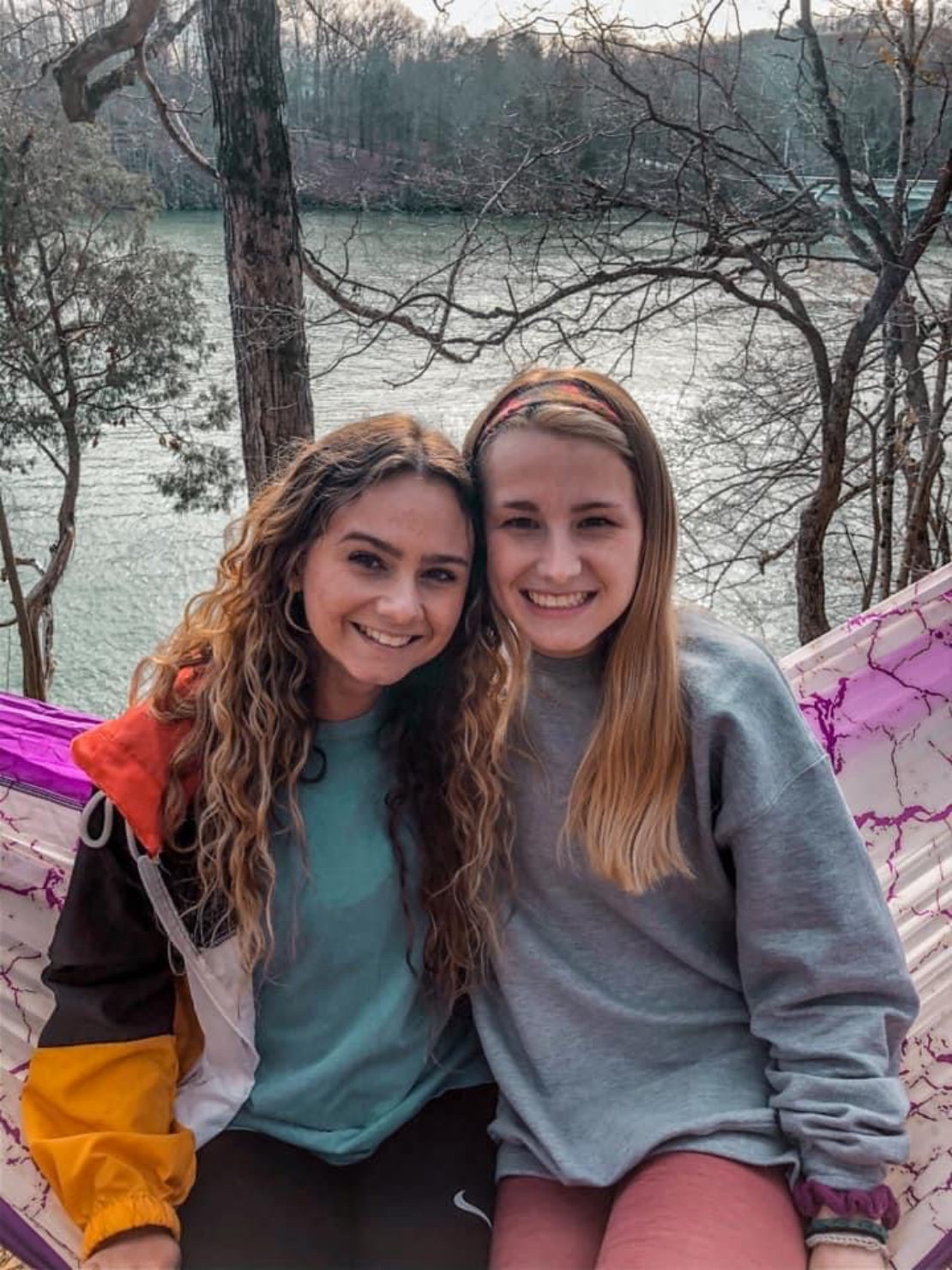 Two young women sit closely together on a colorful hammock outdoors, smiling at the camera. In the background, there are trees and a river, creating a peaceful, natural setting.