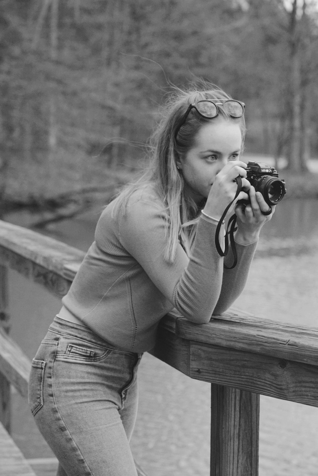 A woman with long hair and glasses on her head leans on a wooden railing, holding a camera to her eye and taking a photo outdoors near water; the image is in black and white.