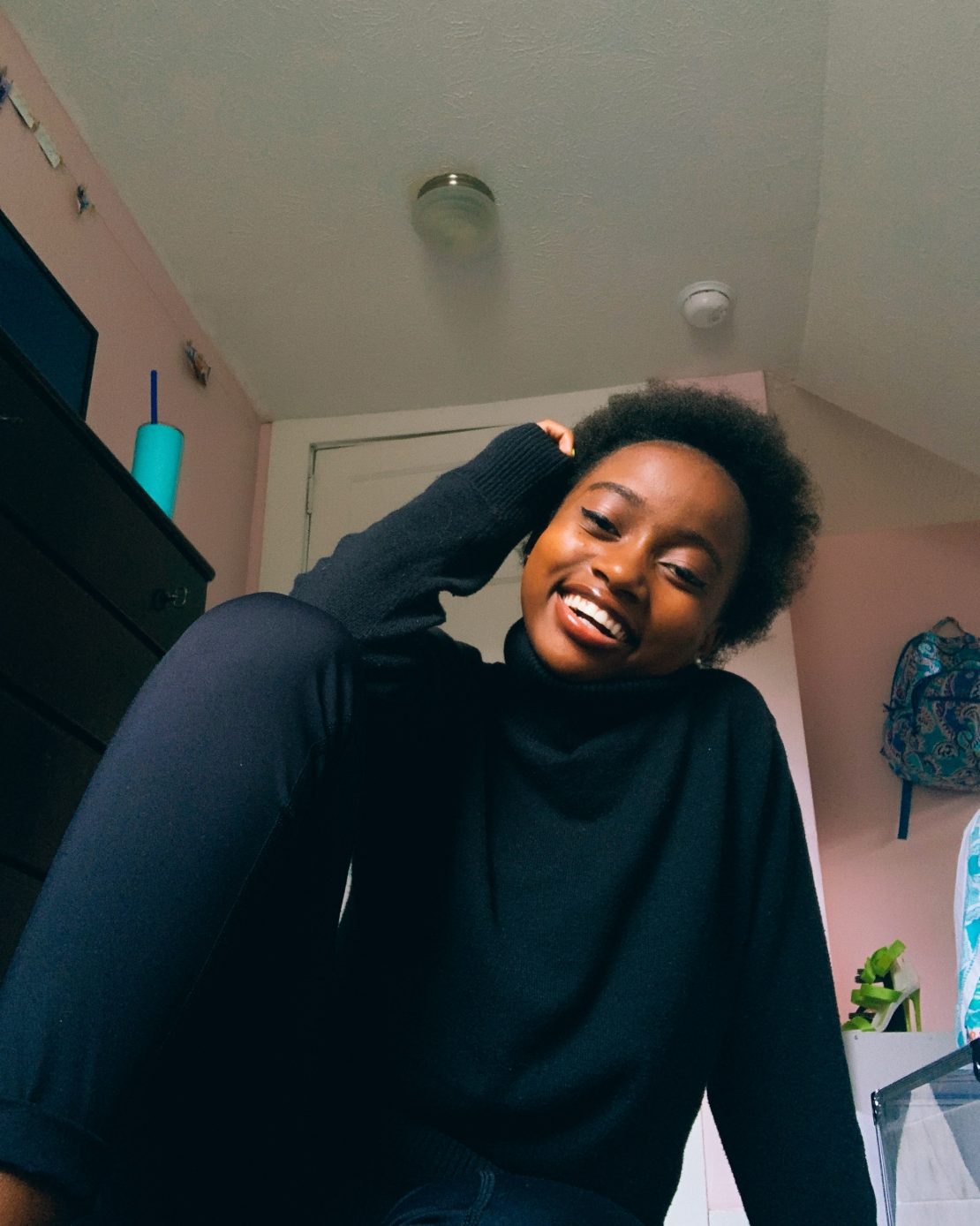 A young woman with short curly hair, wearing a black sweater and pants, sits on the floor smiling with one hand resting on her head in a cozy, pastel-colored room.