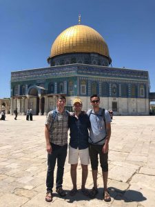 Three men stand smiling arm-in-arm in front of the Dome of the Rock, a gold-domed Islamic shrine in Jerusalem, on a sunny day with a clear blue sky. A few other people are visible in the background.