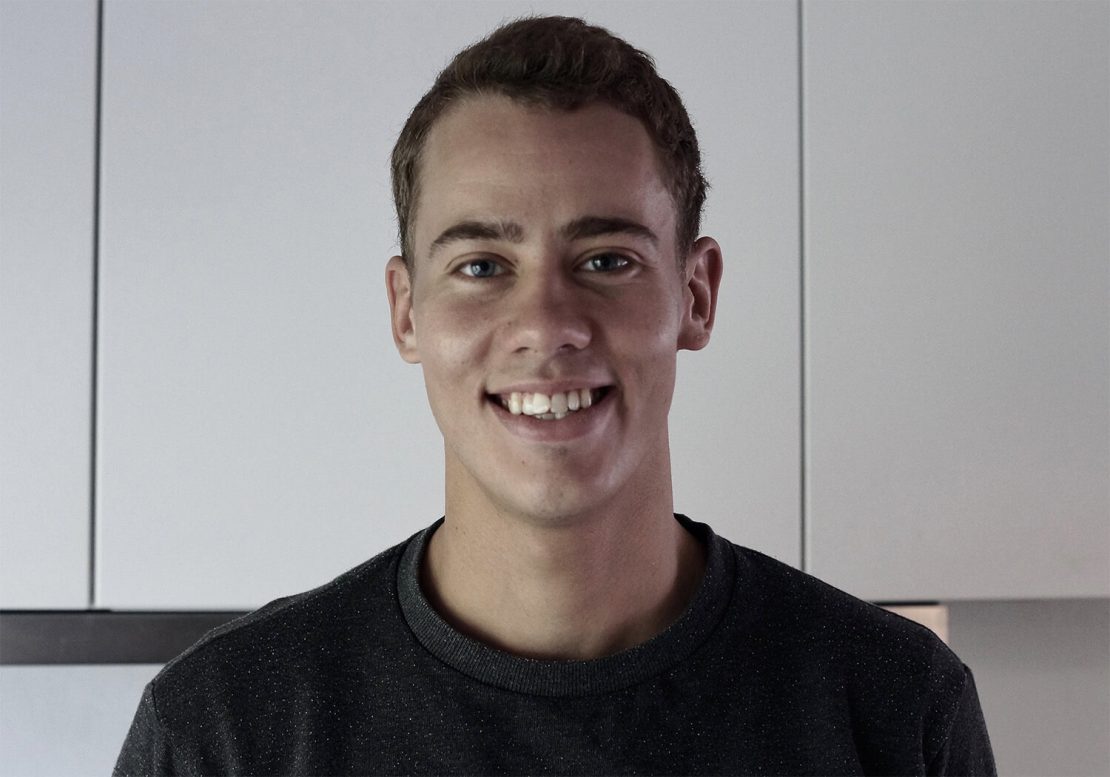 A young man with short brown hair smiles at the camera, standing in front of white cabinets in a modern kitchen. He is wearing a dark-colored sweater.