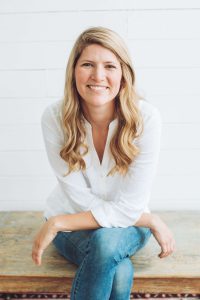 A woman with long blonde hair, wearing a white shirt and blue jeans, sits cross-legged on a wooden bench in front of a white shiplap wall, smiling at the camera.