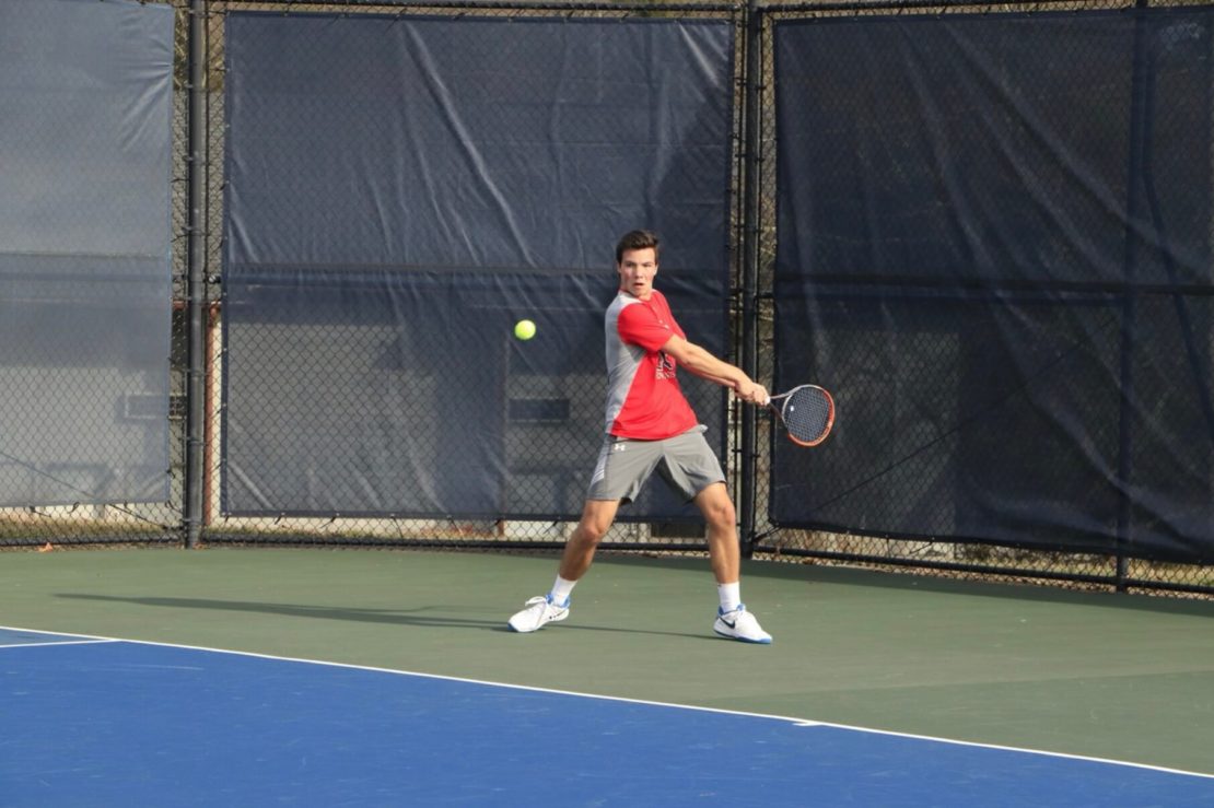 A tennis player in a red shirt and gray shorts prepares to hit a backhand shot on an outdoor court, with a green and blue surface and a chain-link fence in the background.