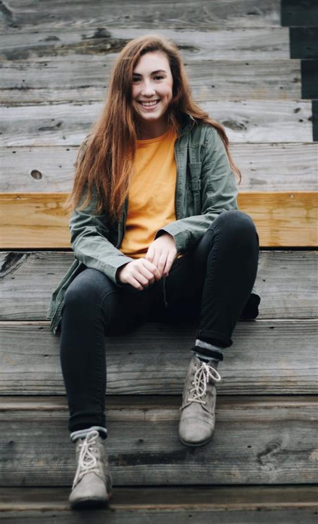A young woman with long brown hair, wearing a yellow shirt, green jacket, black jeans, and gray boots, sits on outdoor wooden steps and smiles at the camera—celebrating her success in earning academic scholarships.