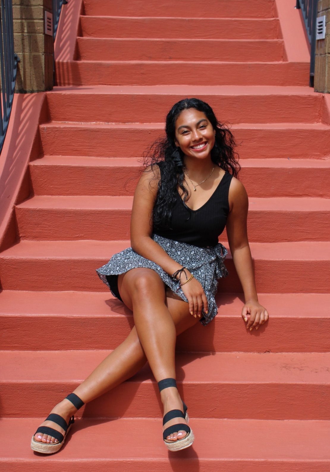 A smiling woman with long dark hair sits on red stairs, wearing a black sleeveless top, a patterned skirt, and black platform sandals. She has one leg extended and her hands rest on her lap and the step beside her.