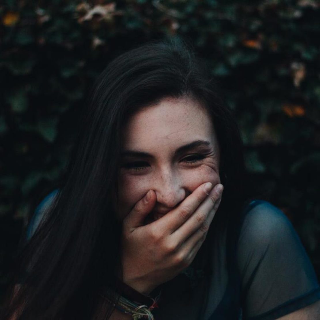 A person with long dark hair smiles and covers their mouth with one hand, standing outdoors in front of a leafy background. The soft, slightly dim lighting gives a contemplative feel, reminiscent of students reflecting on academic scholarships.
