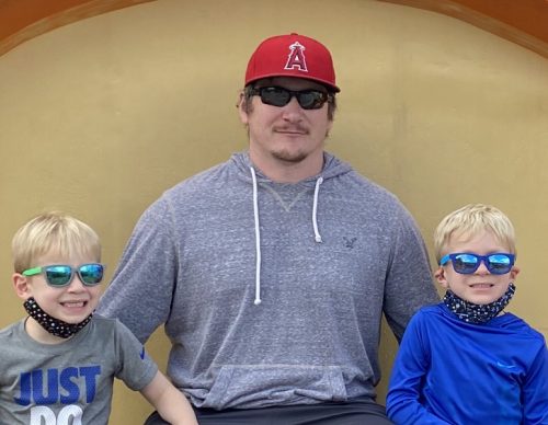 A man in a red baseball cap and gray hoodie sits between two young boys wearing sunglasses and face masks under their chins. The boys are smiling, and all three are seated in front of a yellow background.