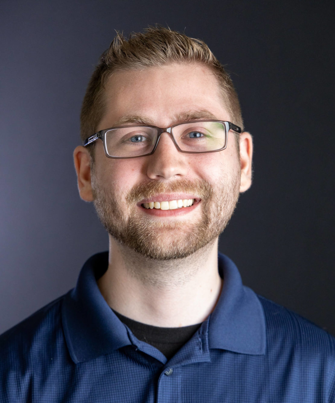 A young man with short light brown hair, glasses, a trimmed beard, and a blue collared shirt smiles in front of a plain dark background.