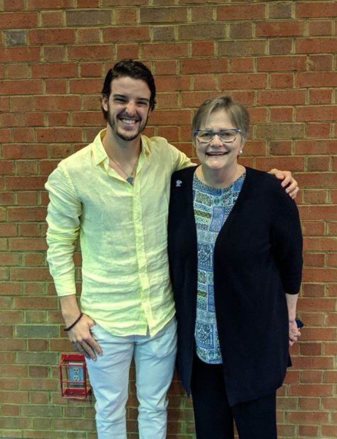 A young man in a light yellow shirt and white pants stands smiling next to an older woman in glasses and a blue patterned top, both in front of a brick wall.