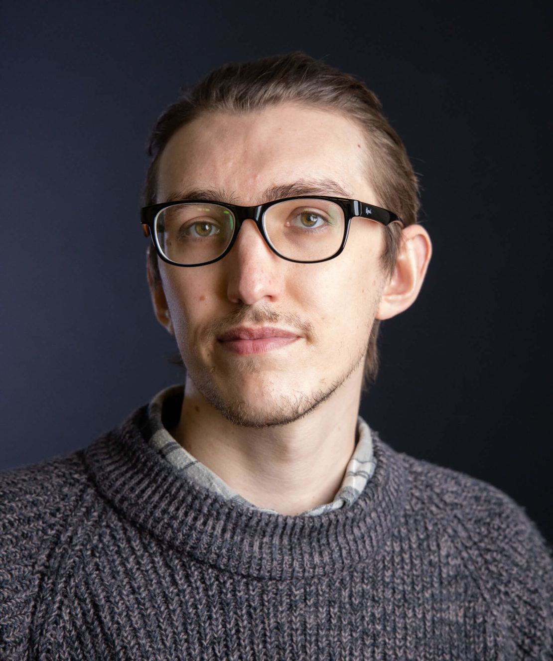 A person with light skin, short brown hair, and glasses is wearing a gray knit sweater over a plaid shirt, looking at the camera with a neutral expression against a dark background.