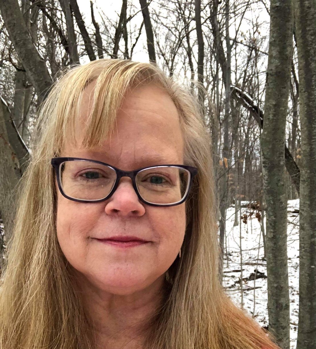 A woman with long blonde hair and glasses smiles softly at the camera while standing outdoors in a snowy, leafless forest.