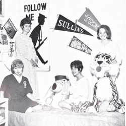 Four young women sit and stand in a dorm room decorated with college pennants, a Follow Me sign, and large stuffed animal toys, including a dog and a tiger. They are smiling and appear relaxed.