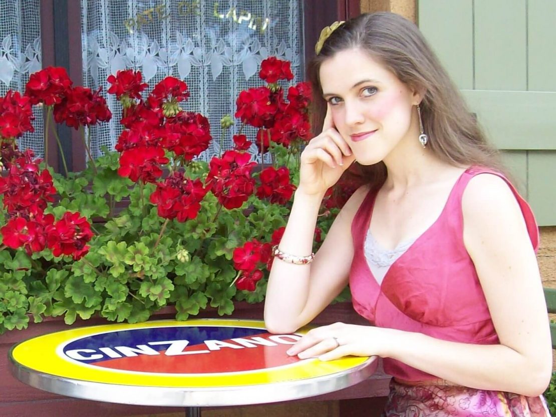 A woman in a sleeveless pink dress sits at a round table with a Cinzano logo in front of a window and vibrant red flowers, smiling slightly and resting her head on one hand.