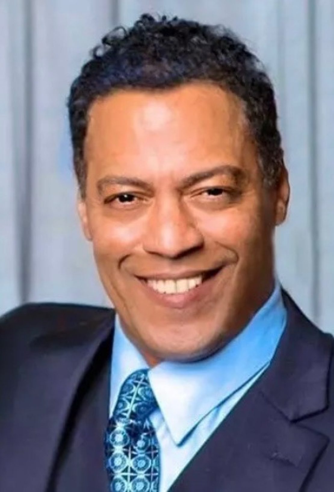 A man wearing a dark suit, light blue shirt, and patterned blue tie smiles at the camera in front of a light-colored background.