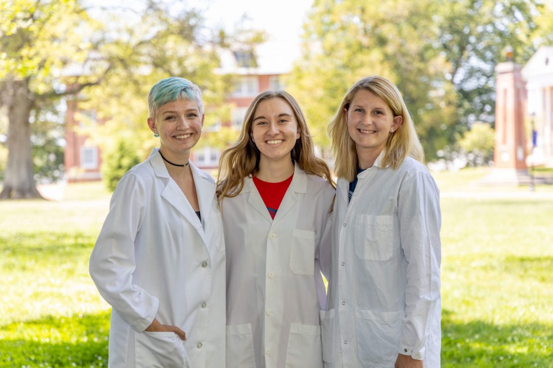 Three young women wearing white lab coats stand outdoors, smiling at the camera. They are surrounded by green grass and trees, with sunlight and buildings visible in the background.