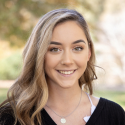 A young woman with long, wavy blonde hair smiles at the camera. She is wearing a black top and a necklace with a round pendant. The background is outdoors and softly blurred with greenery.