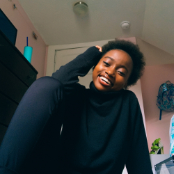 A person wearing a black turtleneck sits indoors, smiling with one hand resting on their head. The room has pink walls, a dresser, a backpack, and a cup with a straw on top.