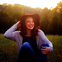 A young woman with wavy brown hair sits in a grassy field at sunset, smiling widely. She wears a striped shirt, denim overalls, and a wide-brimmed red hat, with trees and golden sunlight in the background.